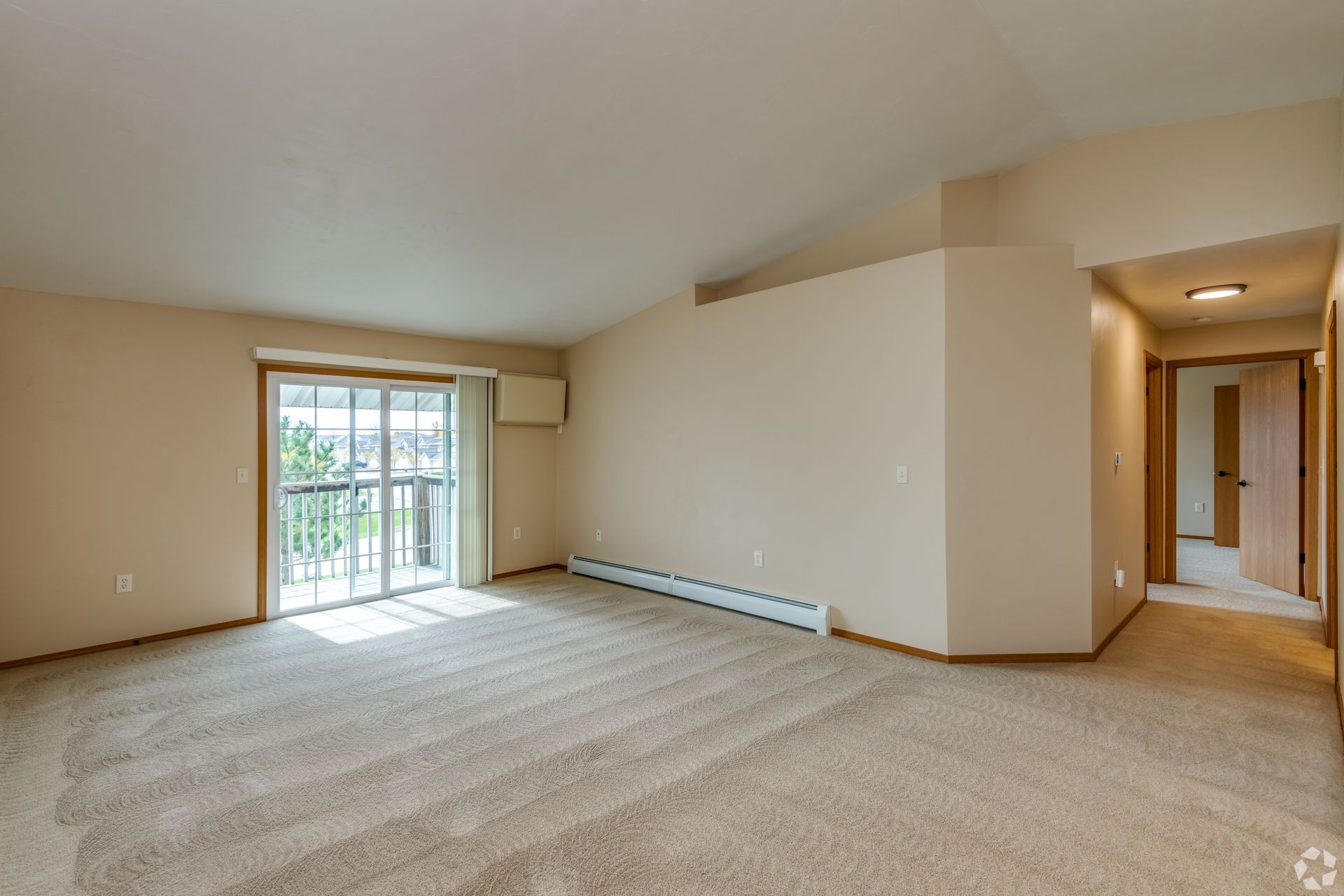 Empty carpeted living room with sliding glass door, beige walls, and hallway opening to the right