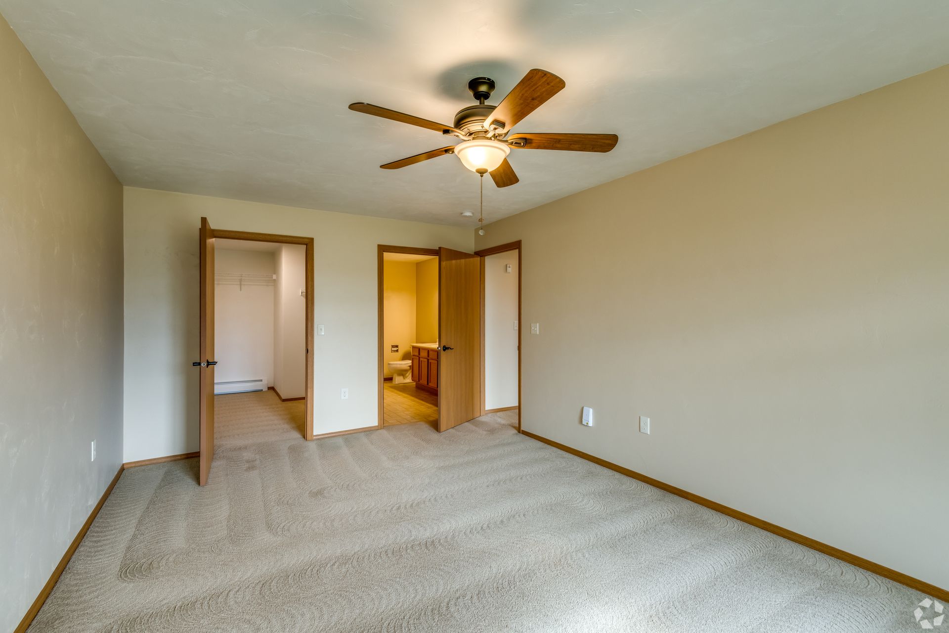 Empty beige carpeted bedroom with ceiling fan, two doorways, and attached bathroom visible