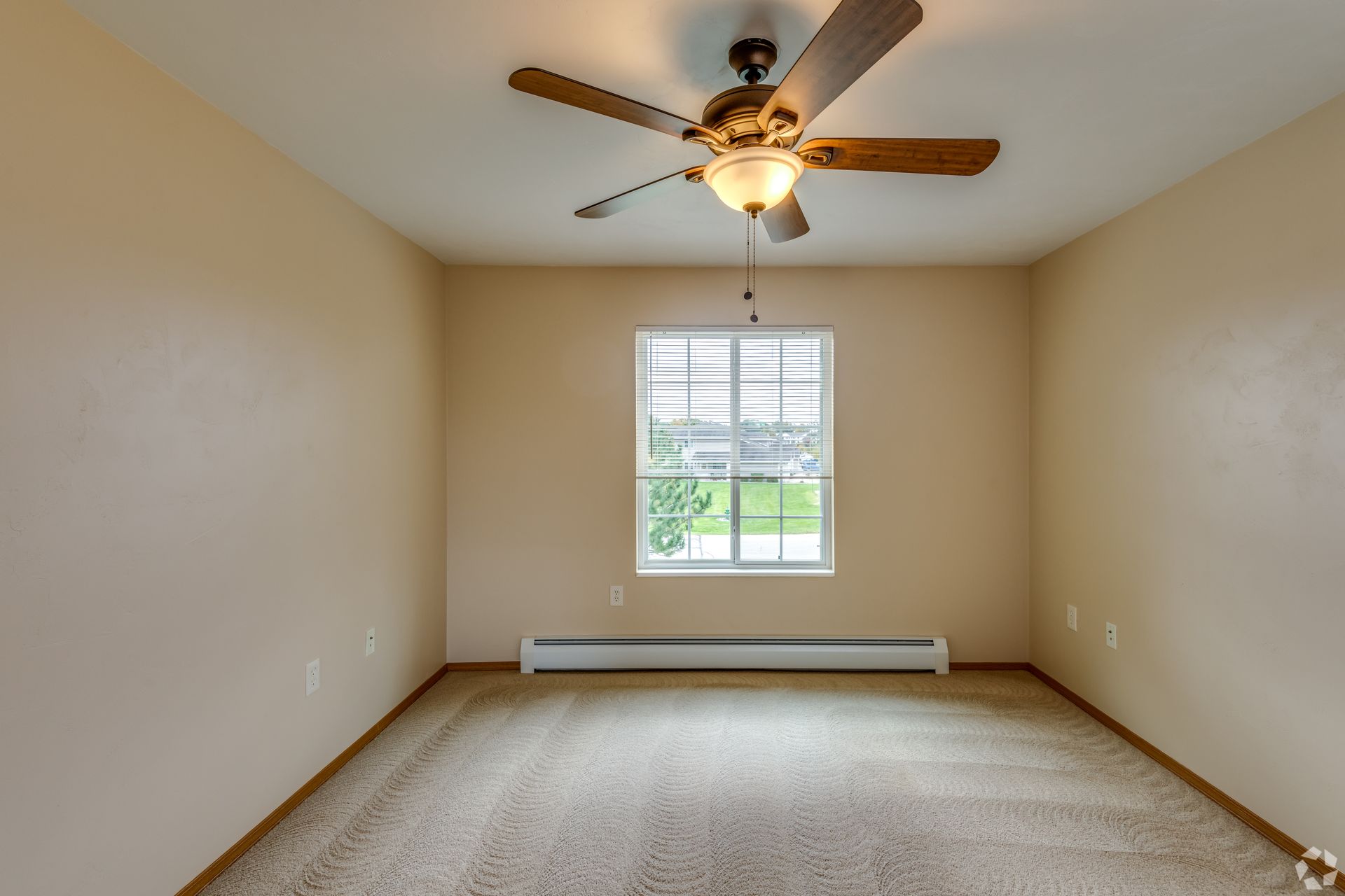 Empty beige bedroom with carpet, window, baseboard heater, and ceiling fan light.