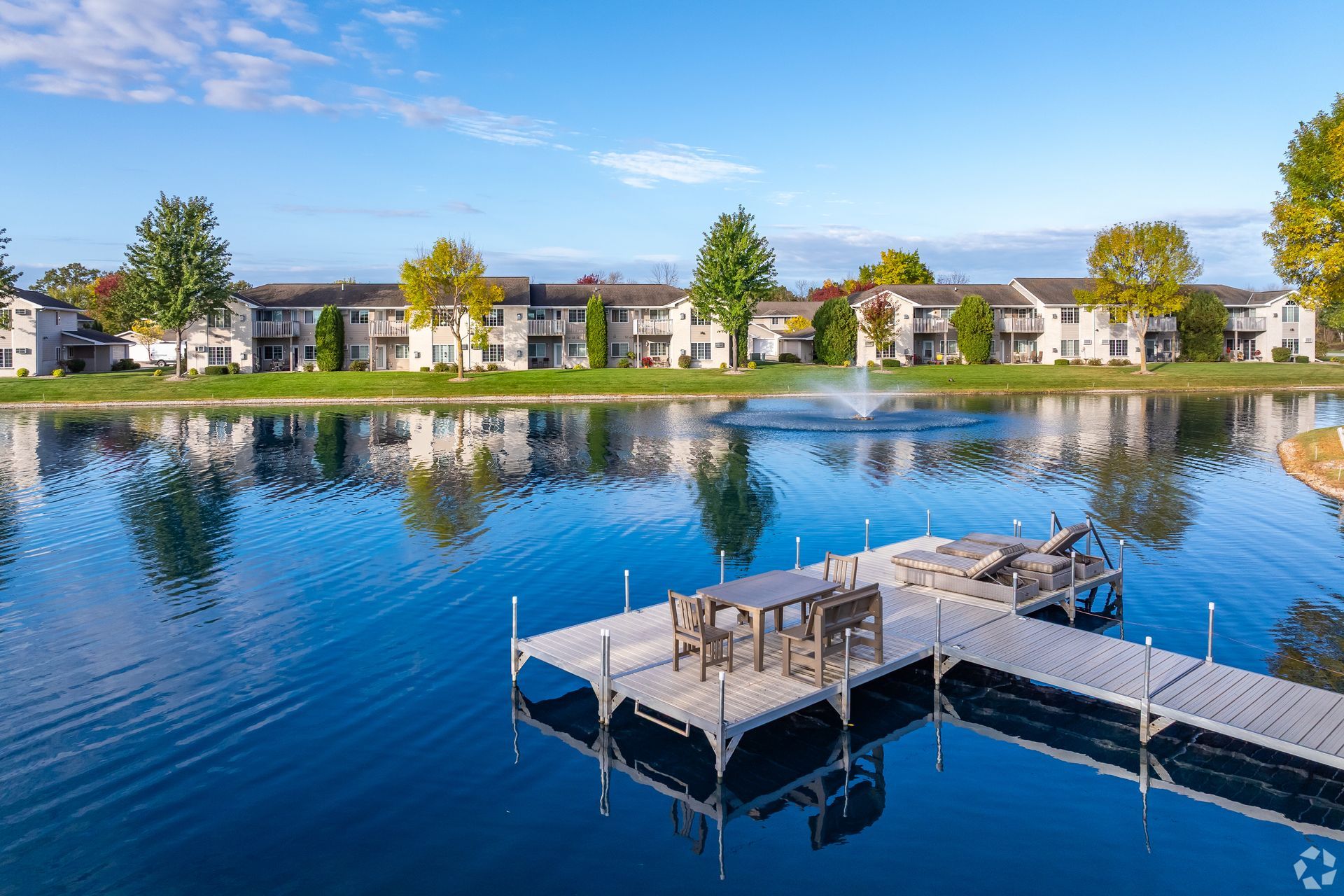 Dock extending into a calm blue lake with waterfront homes and trees reflected in the water.