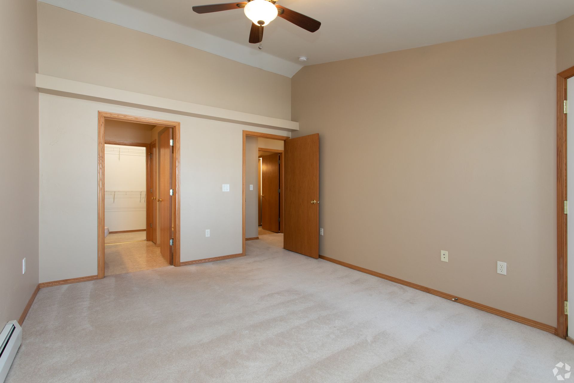 Empty beige bedroom with carpet, ceiling fan, and two open doorways leading to a hallway and closet.