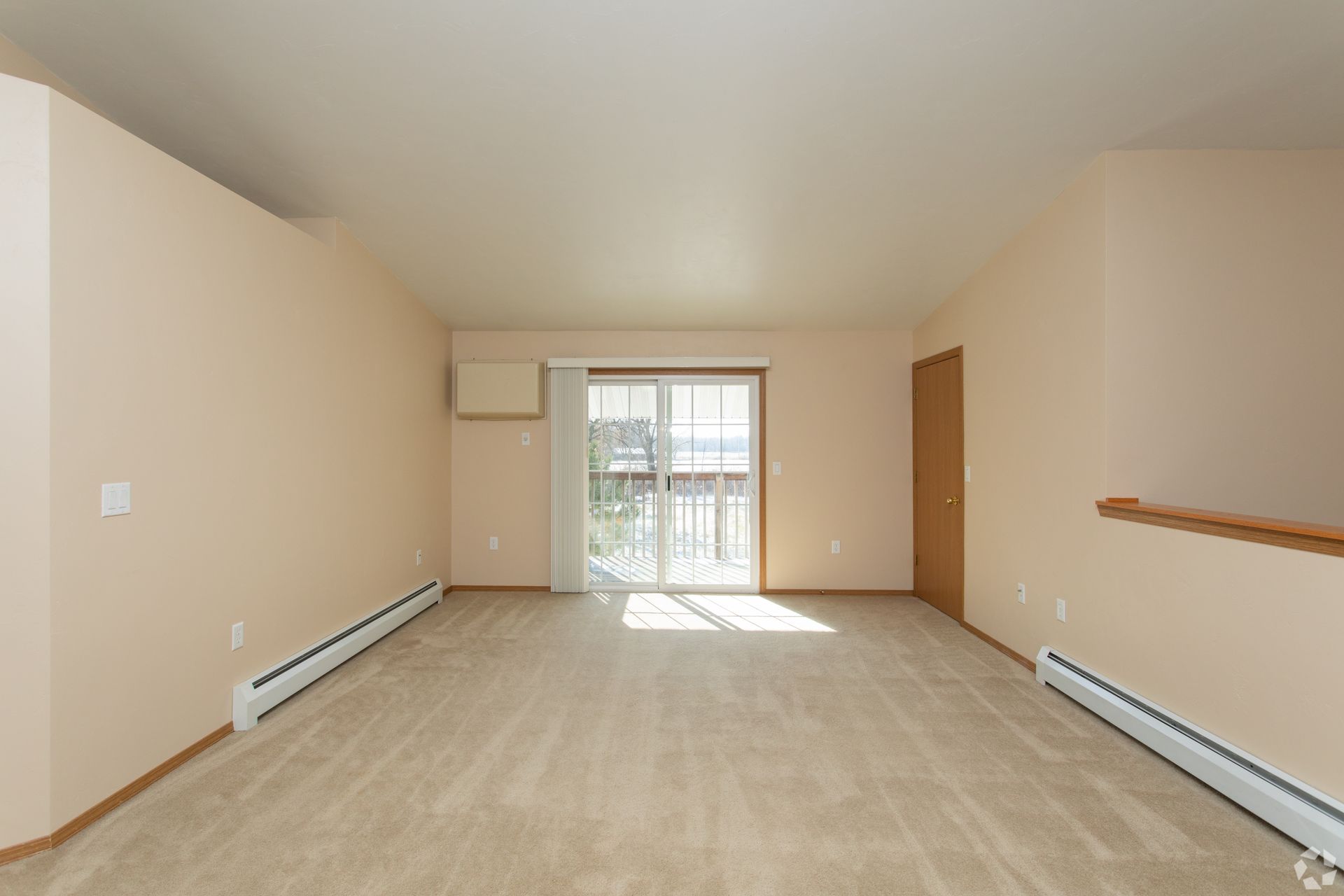Empty beige living room with carpet, wall heater, handrail, and sliding glass doors to a balcony.