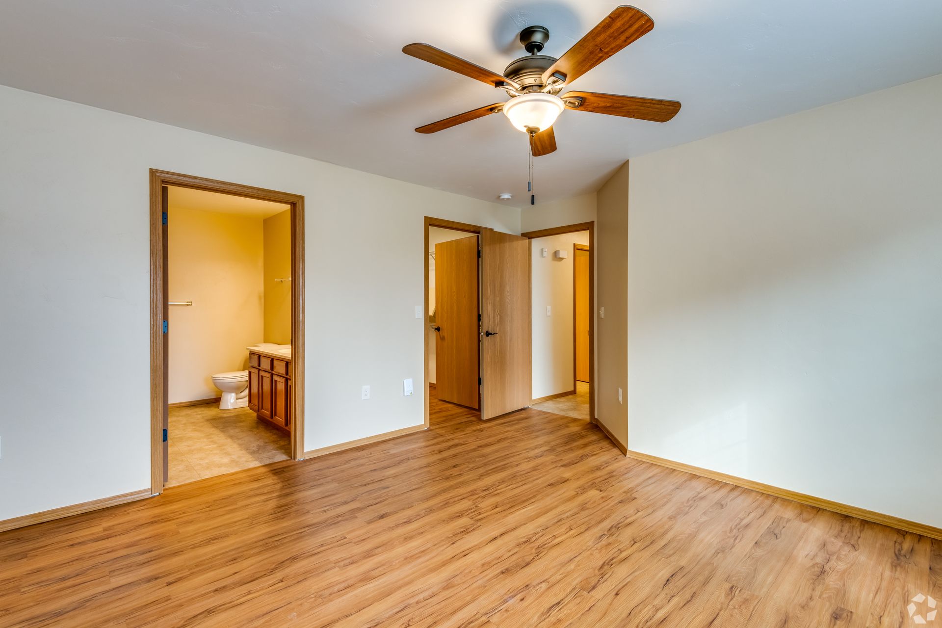 Empty beige bedroom with wood floor, ceiling fan, and open doors to a bathroom and closet