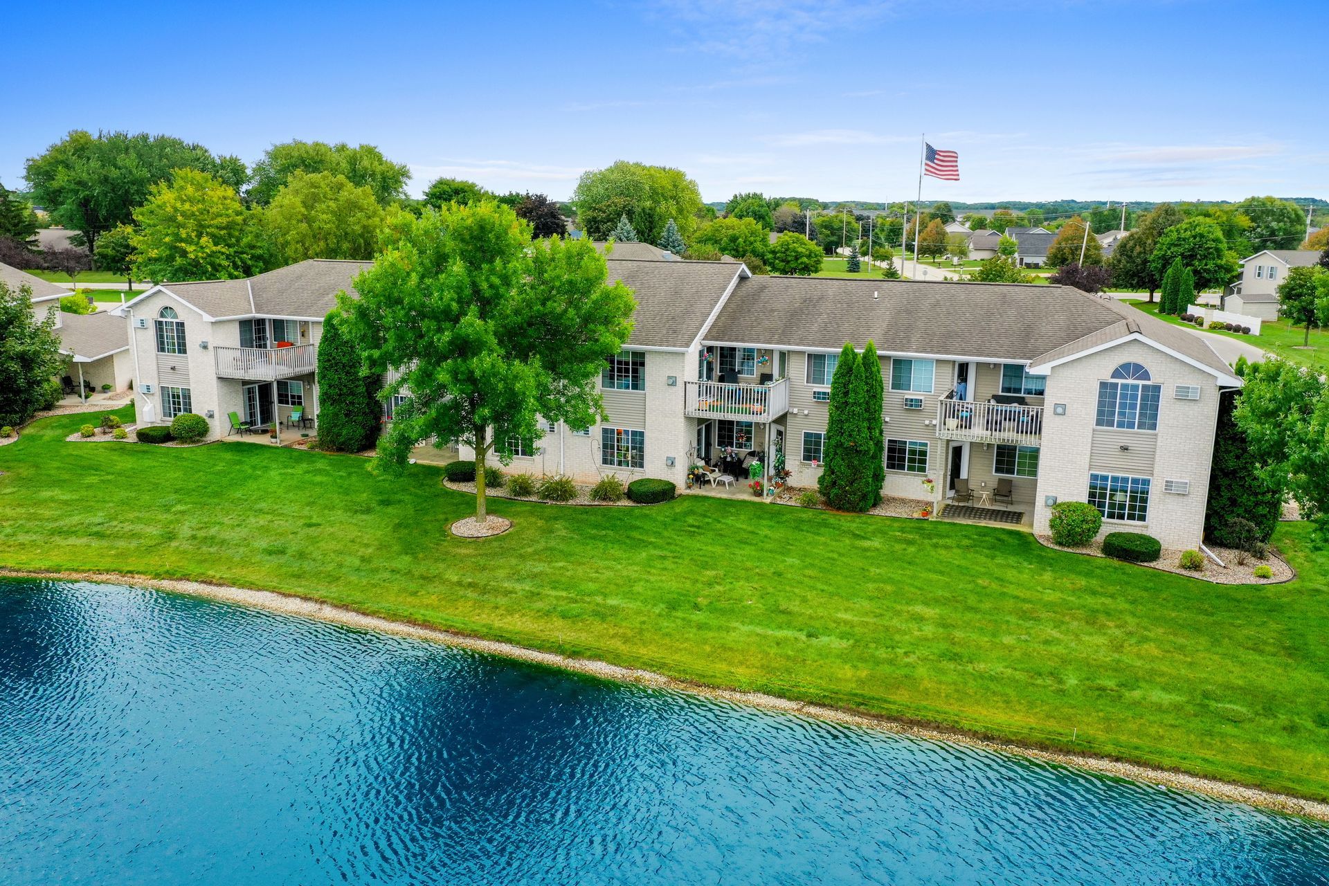 Lakeside apartment buildings with green lawns, trees, and a small American flag under a blue sky
