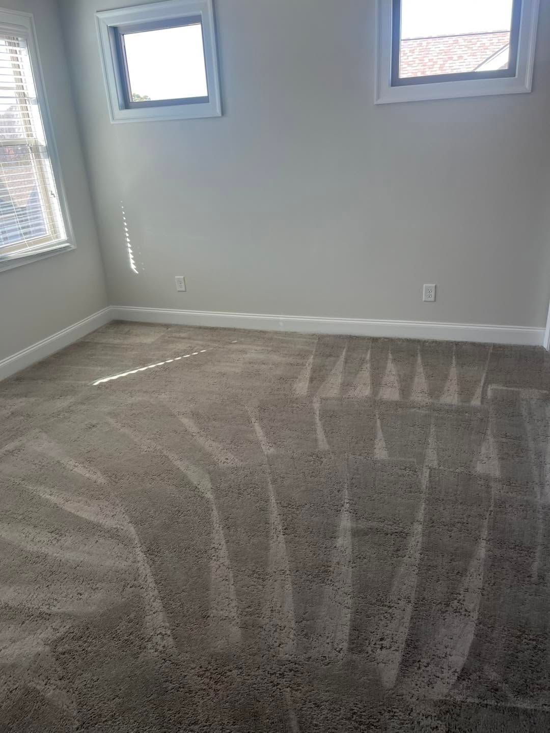 Empty carpeted room with gray walls, two small windows, and visible vacuum marks on the floor.