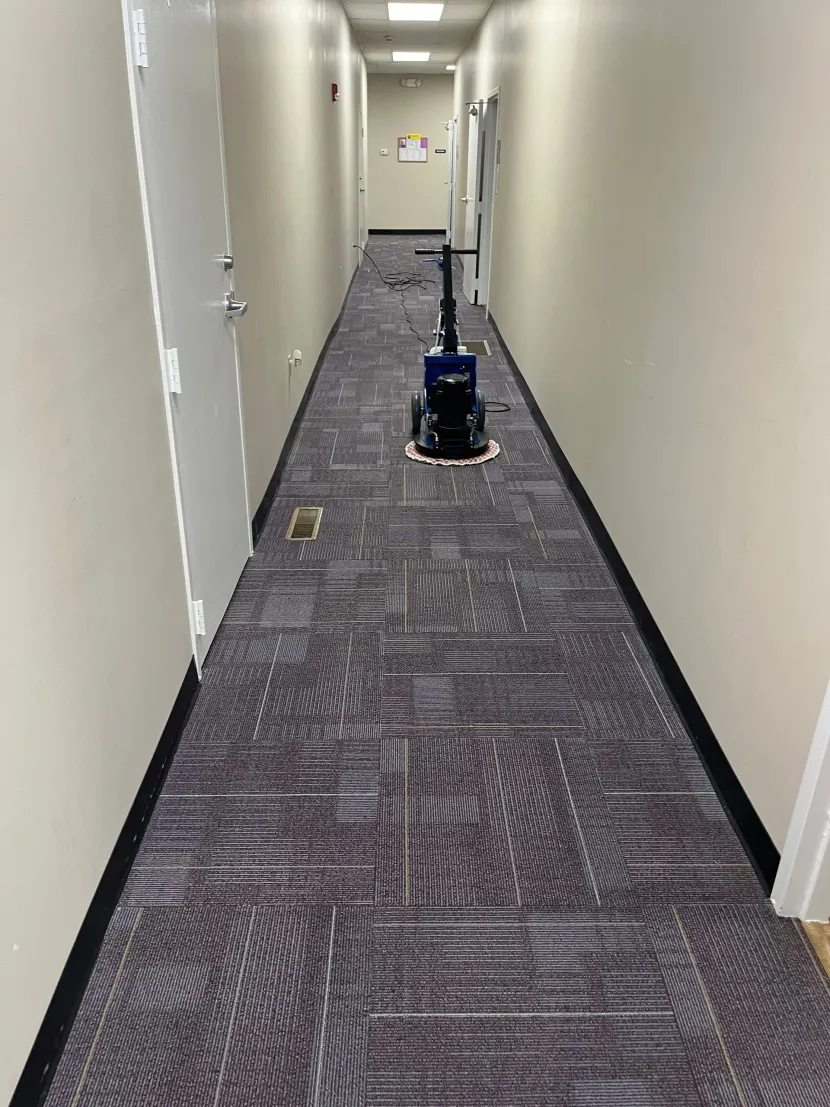 Carpeted hallway with a floor-cleaning robot in the center, beige walls and office doors lining both sides.
