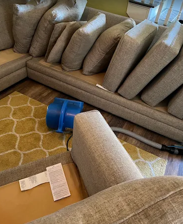 Gray couch with multiple cushions, blue floor fan, and tipped-over chair on a yellow patterned rug.