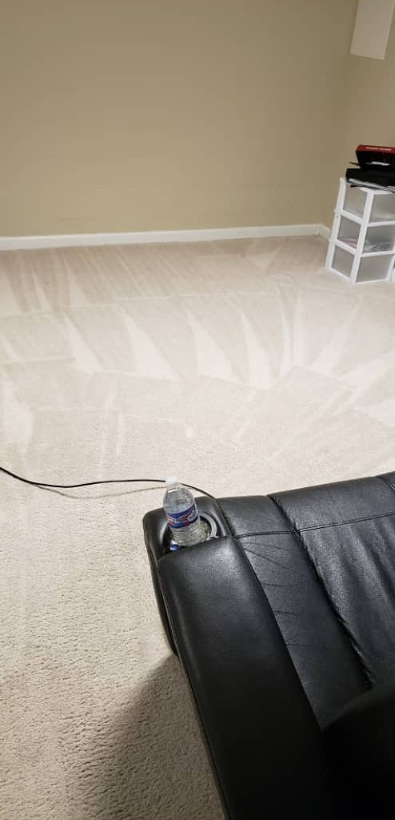 Empty beige carpeted room with black leather chair corner and a white storage bin in the back right.