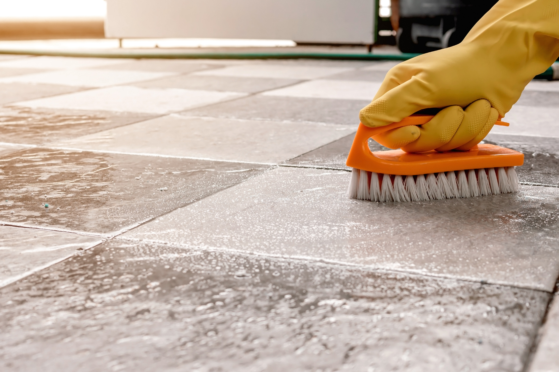 Gloved hand scrubbing a wet tiled floor with an orange brush.