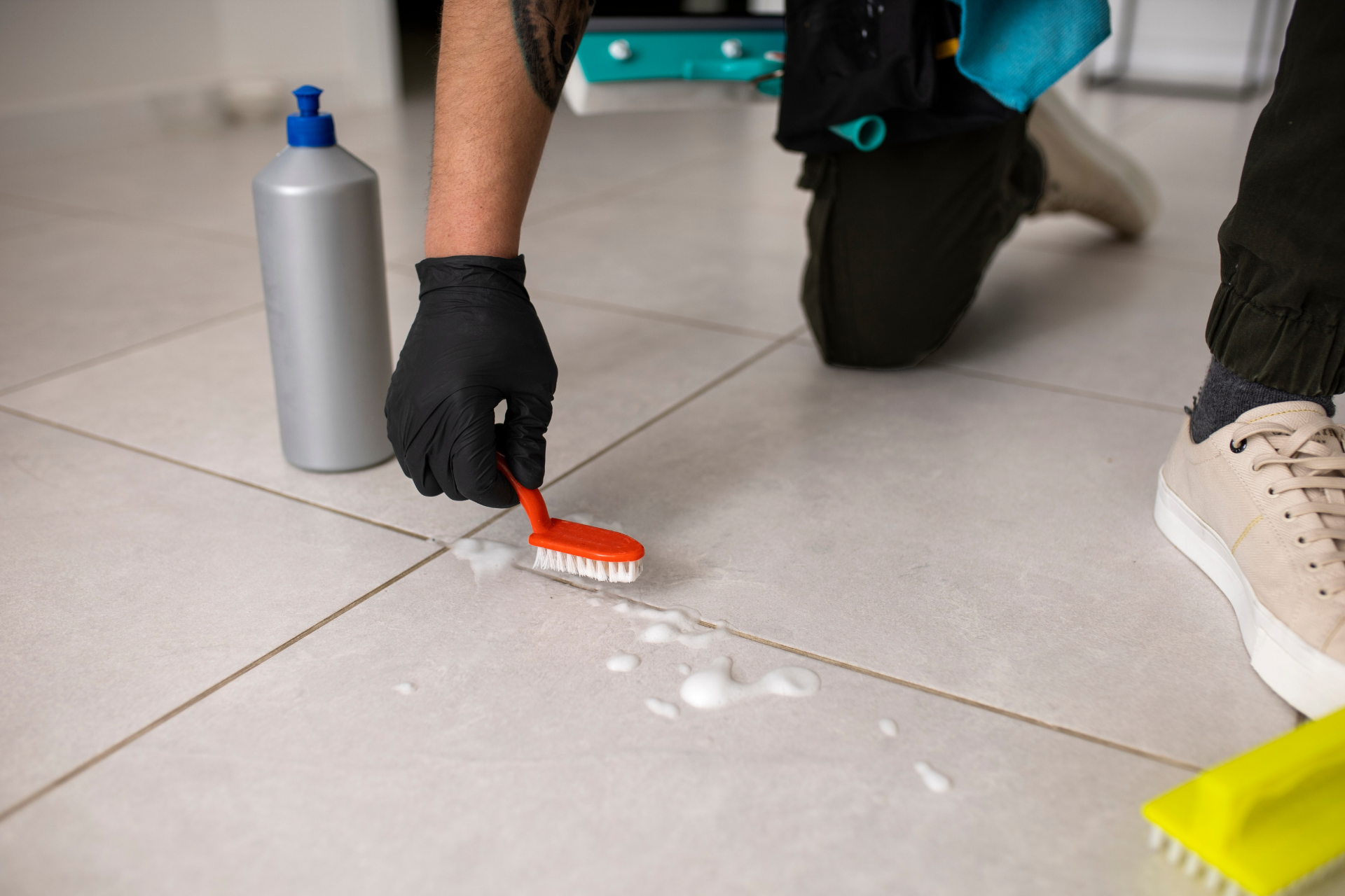 Person cleaning a tile floor with a brush beside a bottle of cleaner and a white spill