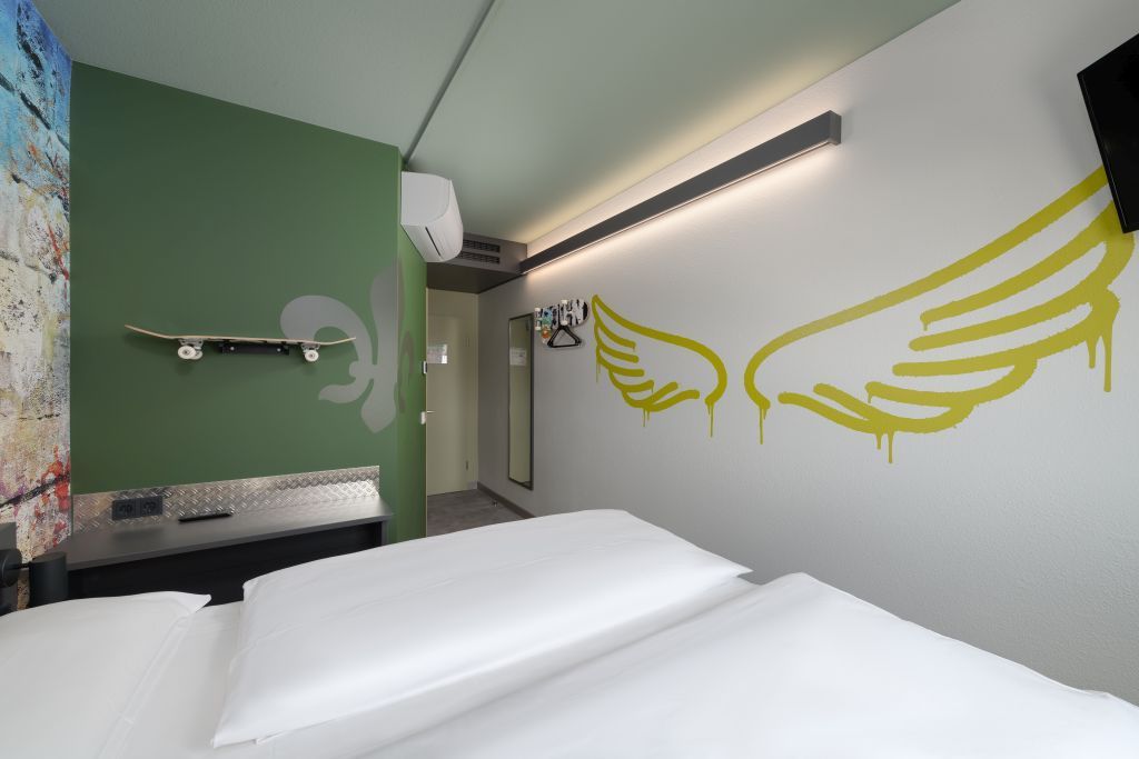 Kleines minimalistisches Schlafzimmer mit weißem Bett, grüner Wand und gelbem Flügelmotiv an der Seitenwand