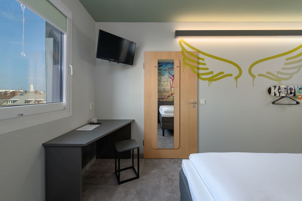 Hotelzimmer mit Bett, Schreibtisch, Wandfernseher, Spiegel und Fenster mit Stadtblick.