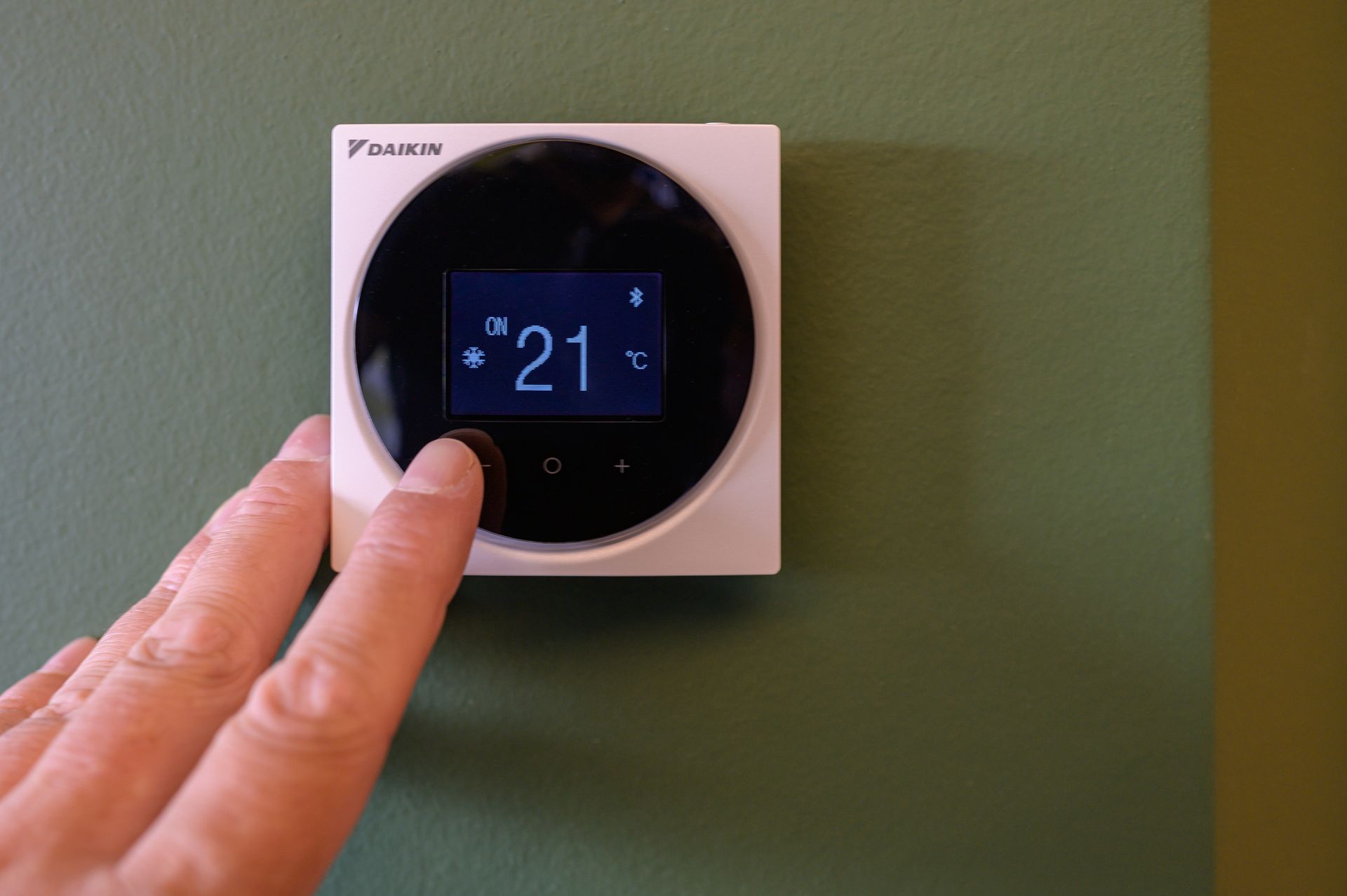 Ein digitaler Thermostat an einer grünen Wand wird manuell eingestellt und zeigt 21° an. Klimaanlage in jedem Zimmer.