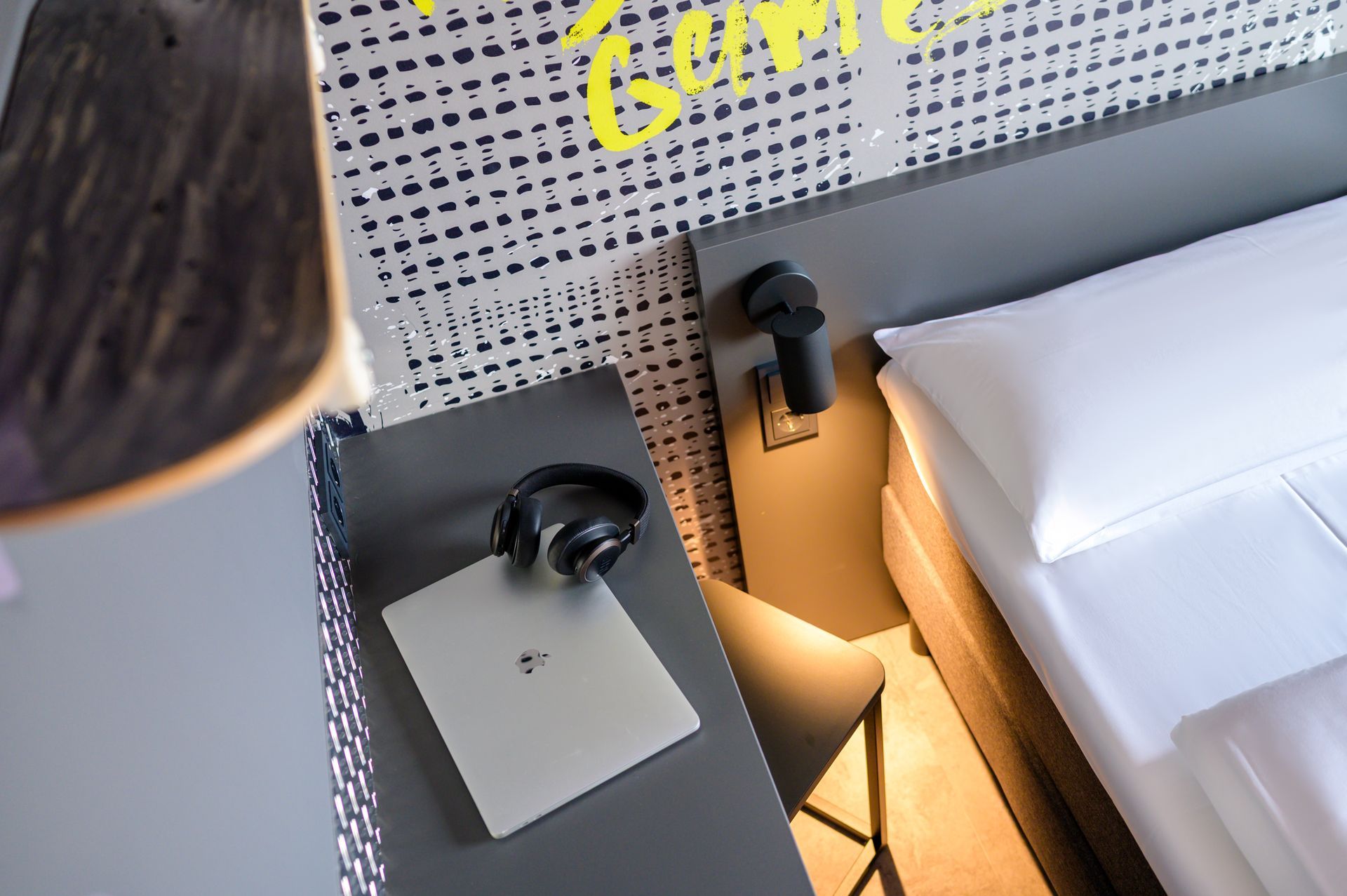 Hotelnachttisch mit Kopfhörern, Laptop, Lampe und einem Bett mit weißer Bettwäsche.