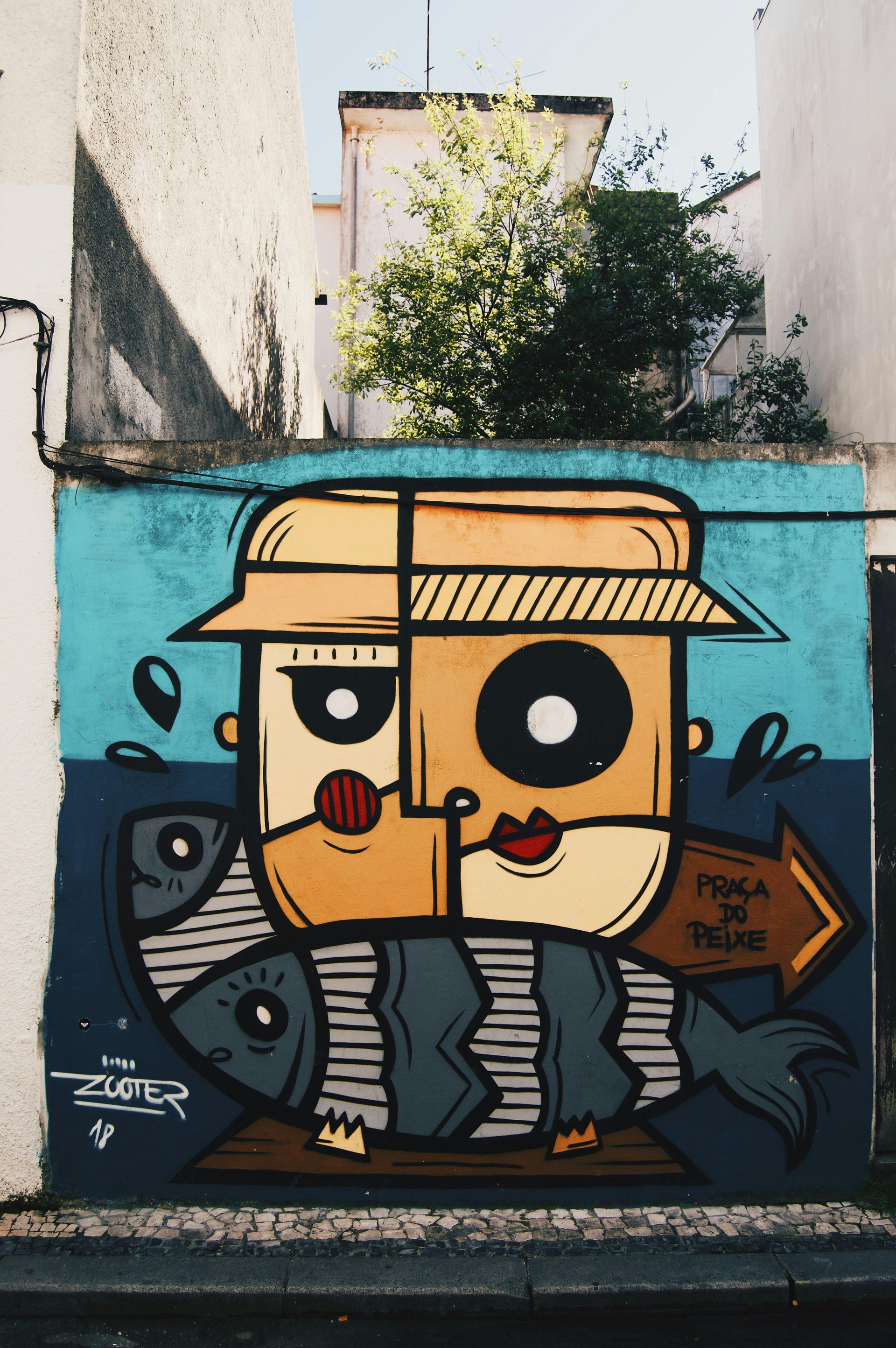 Graffiti-Comicfigur mit Strohhut an einer blauen Wand zwischen Gassengebäuden