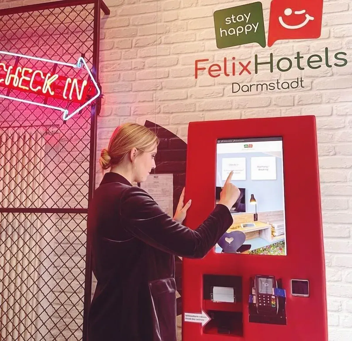 Eine Frau benutzt einen Selbsteincheckautomaten im Felix Hotels Darmstadt unter einem pinkfarbenen „CHECK IN“-Schild.