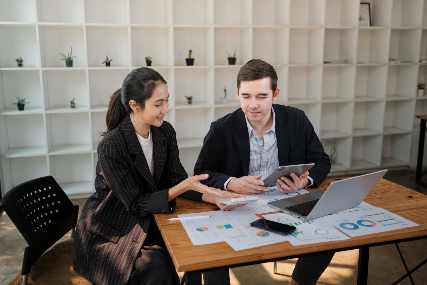 Two coworkers discussing charts at a table with a laptop and tablet in a bright office