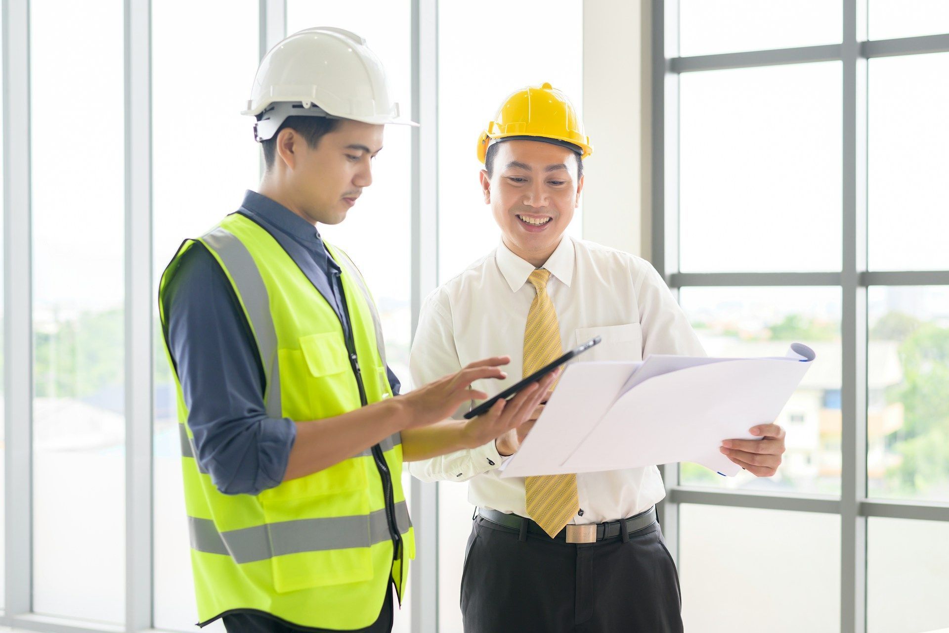 Two engineers in hard hats reviewing plans on a tablet in a bright office by large windows