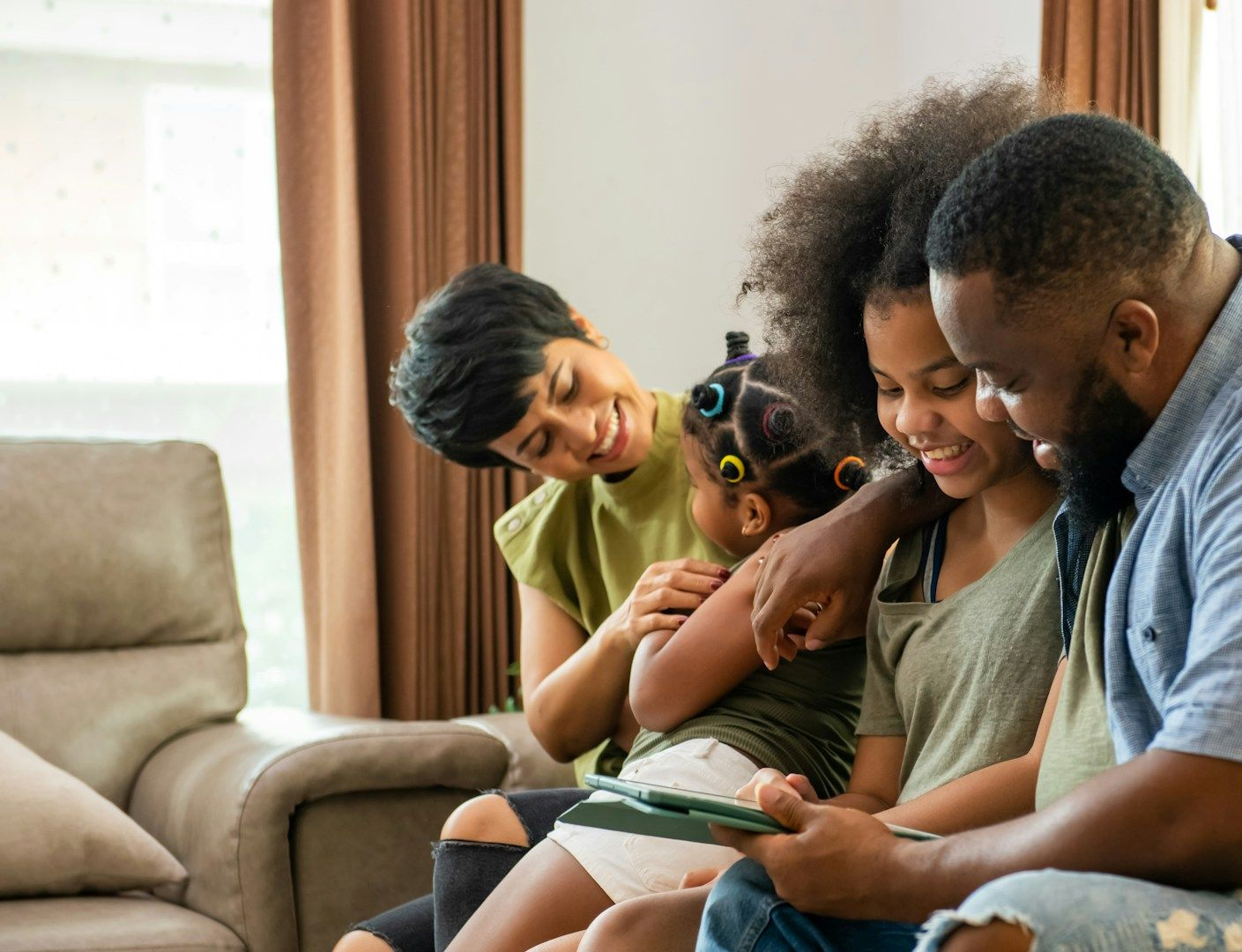 Family sitting on a couch, smiling and looking at a tablet together in a bright living room