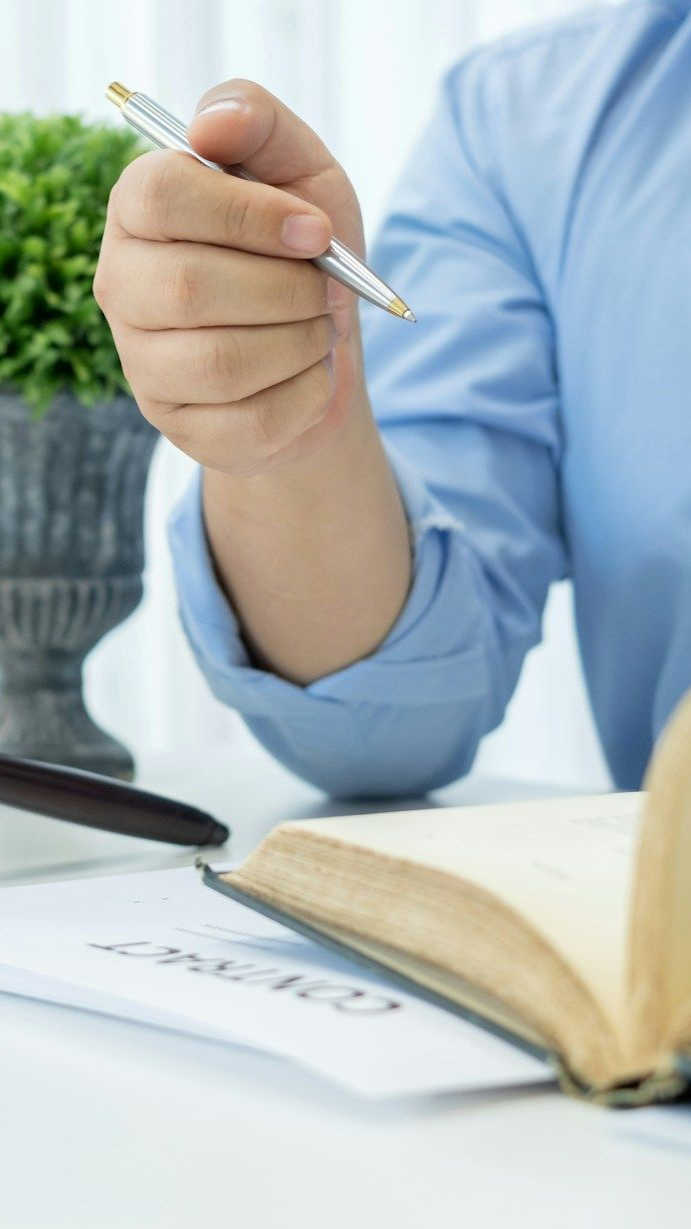 Close-up of a hand holding a pen above an open notebook on a desk, with a laptop nearby.