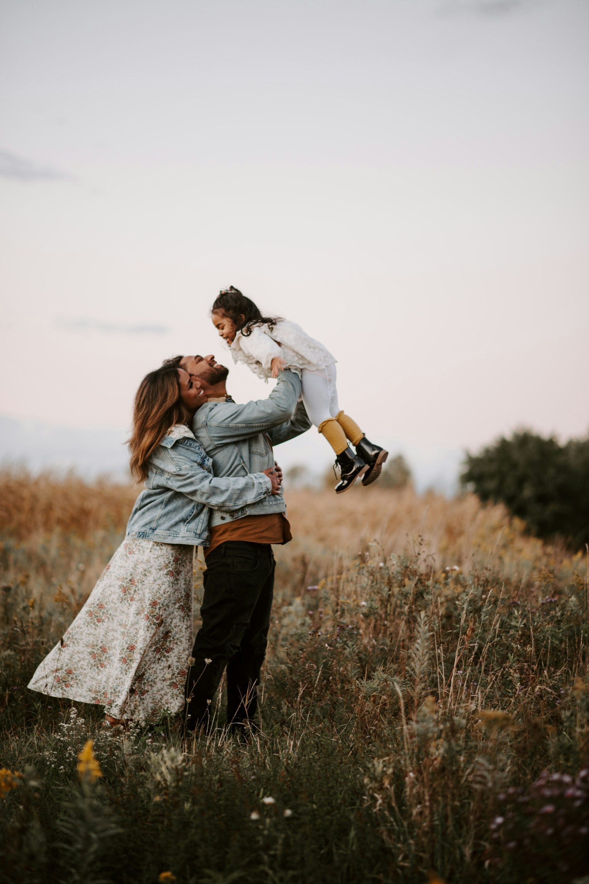 Two people in a grassy field lifting a child in white, outdoors at dusk
