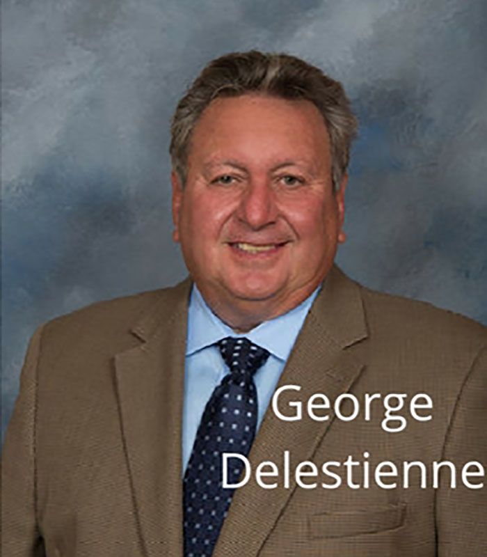 Portrait of George Delestienne in a tan suit and blue tie against a gray studio backdrop
