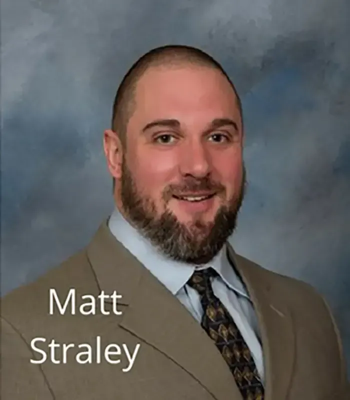 Studio portrait of Matt Straley in tan suit and patterned tie against a gray-blue backdrop
