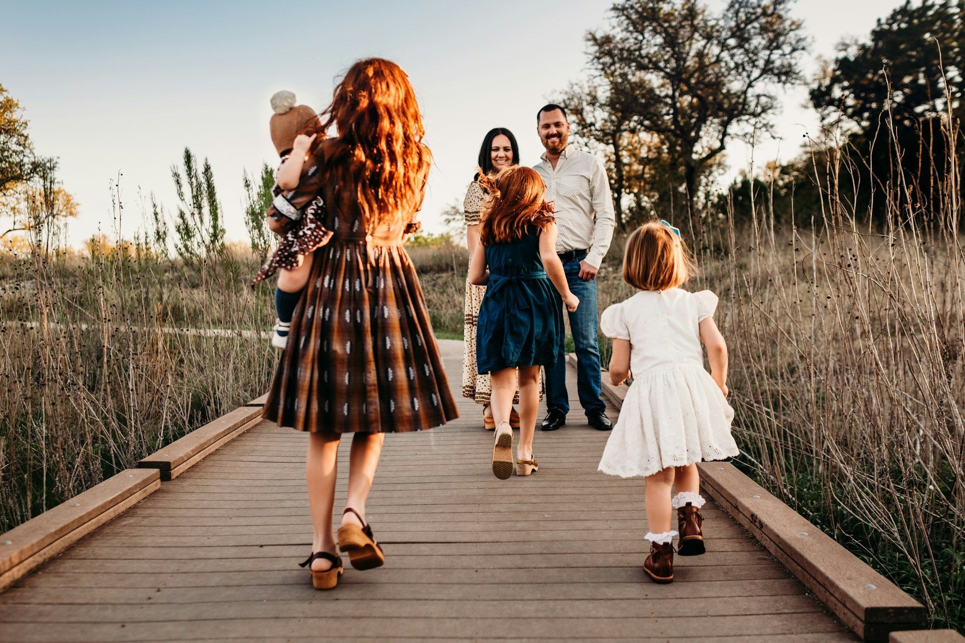 Four people walking along a wooden path through tall grass at sunset