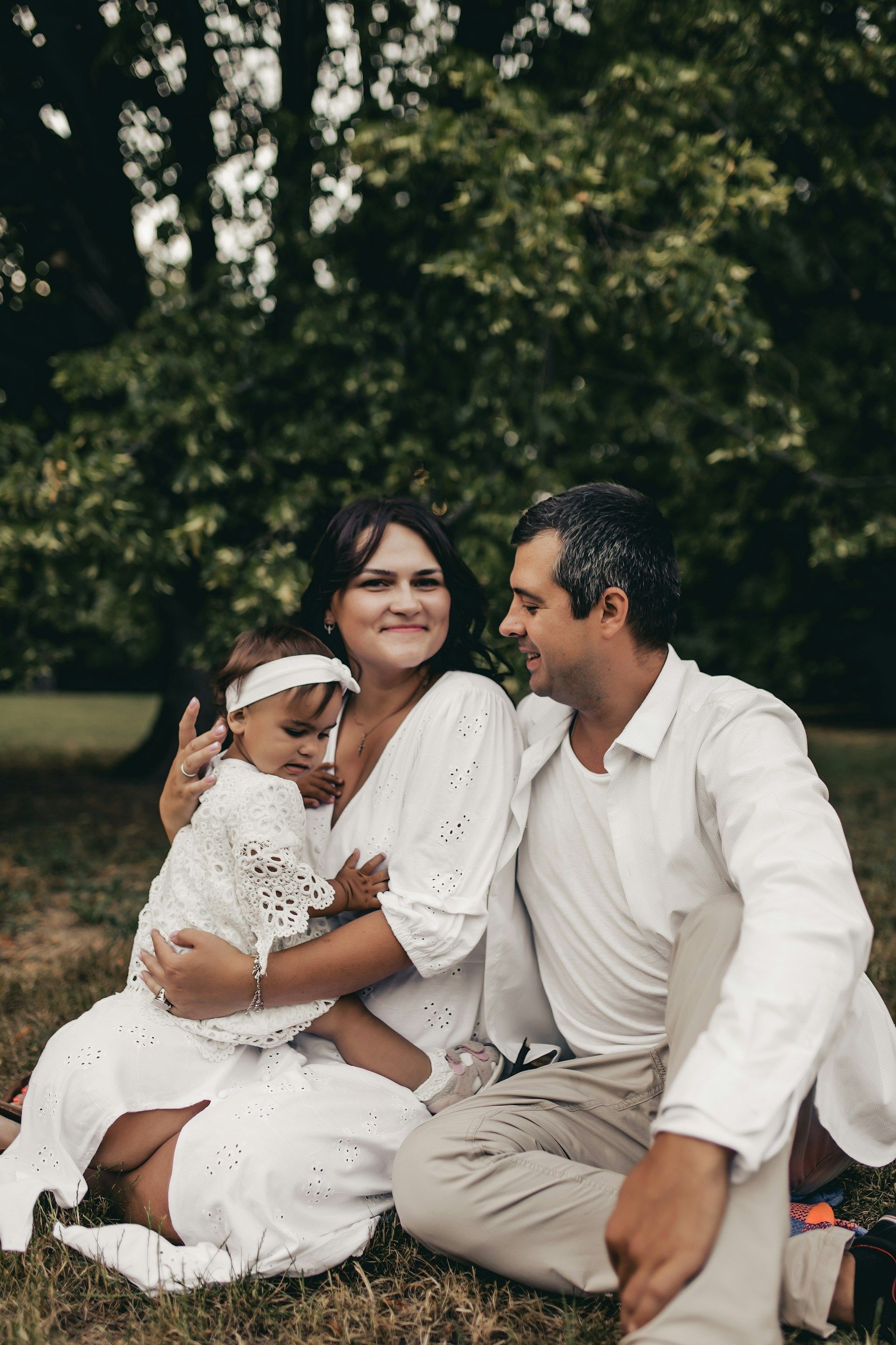 Three people in white clothing sit on grass outdoors, smiling and embracing in a leafy park.