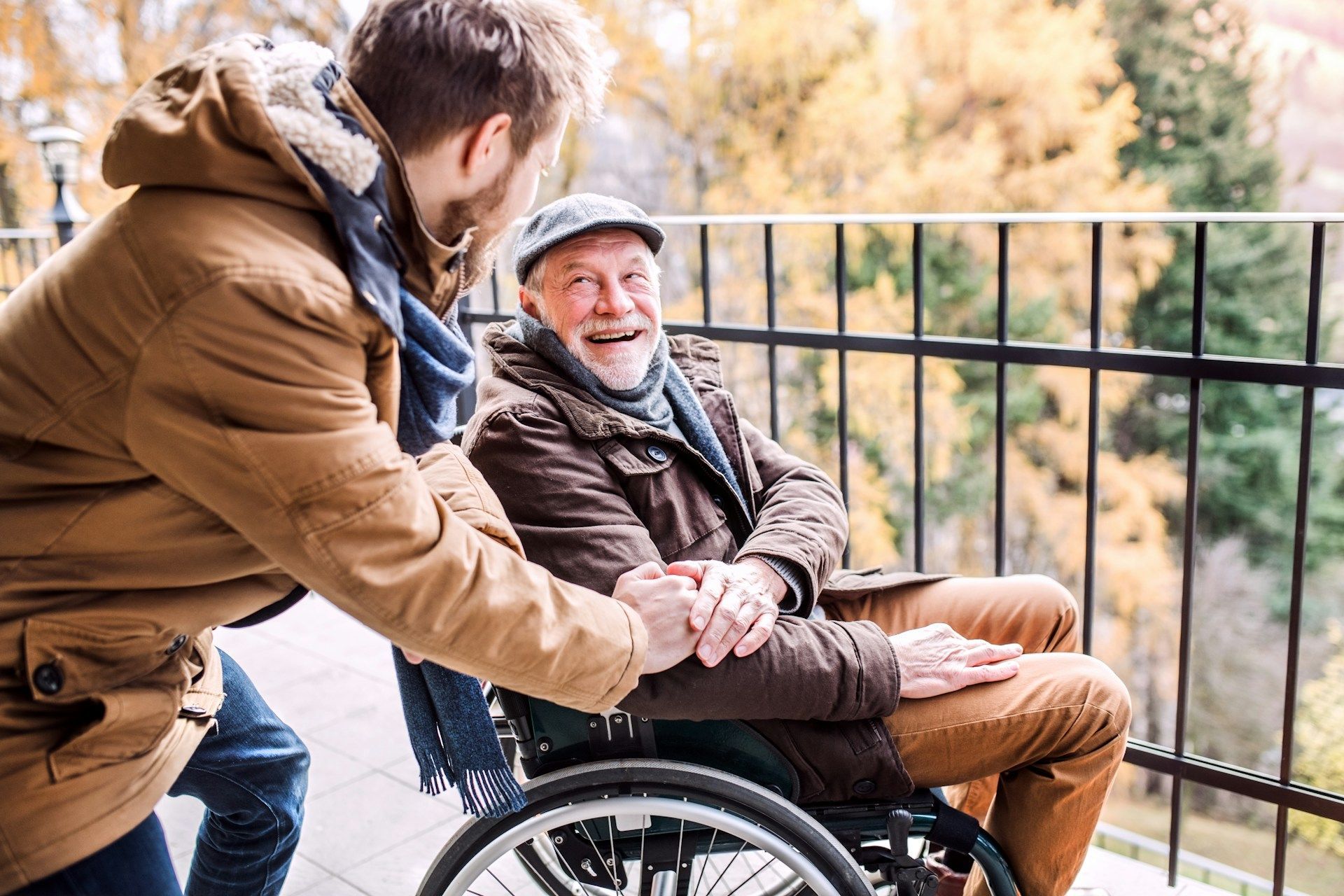 Person in a wheelchair smiles while being pushed outdoors on a balcony in autumn.