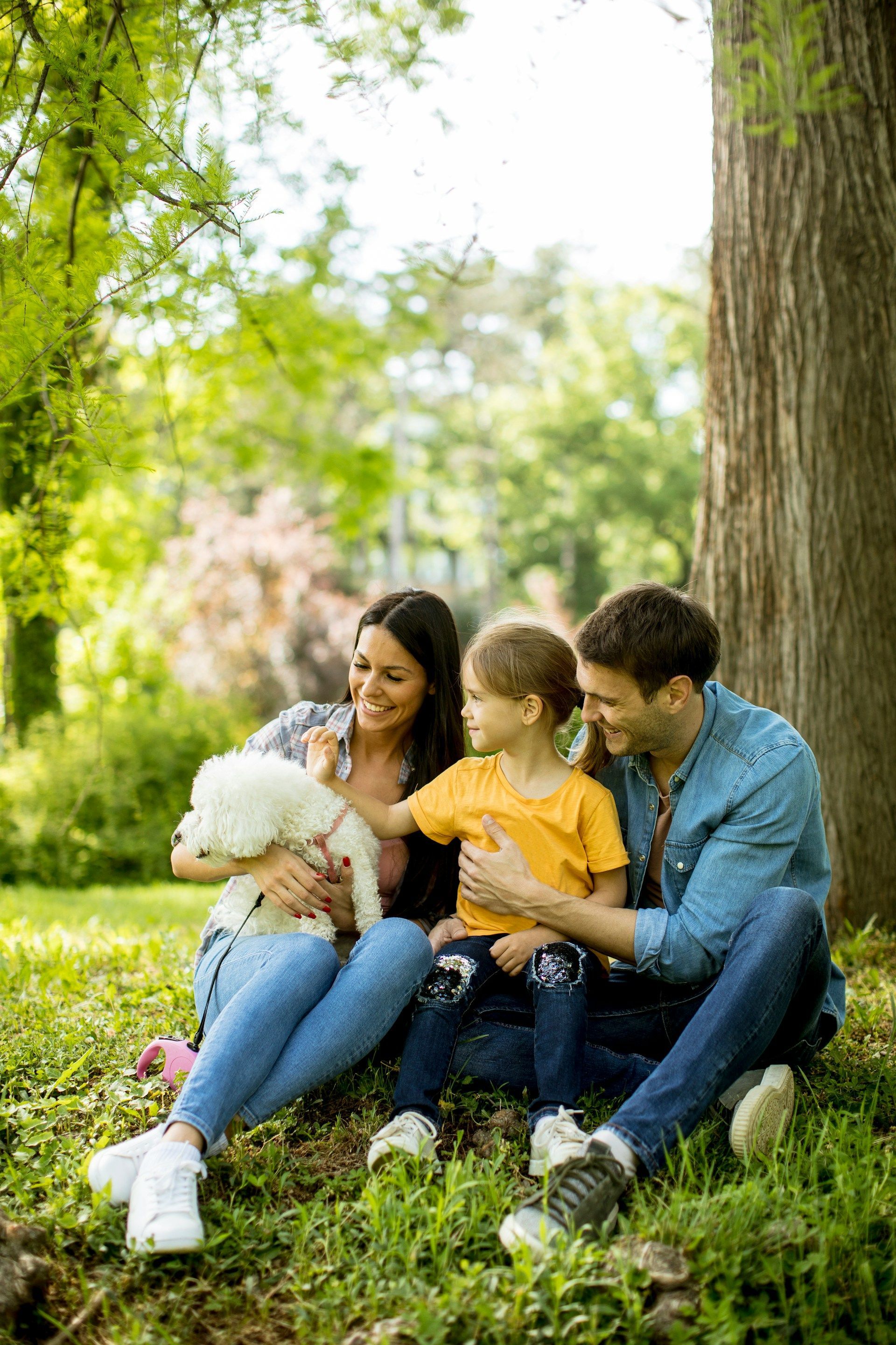 Family sitting on grass in a sunny park, smiling and petting a small white dog under trees