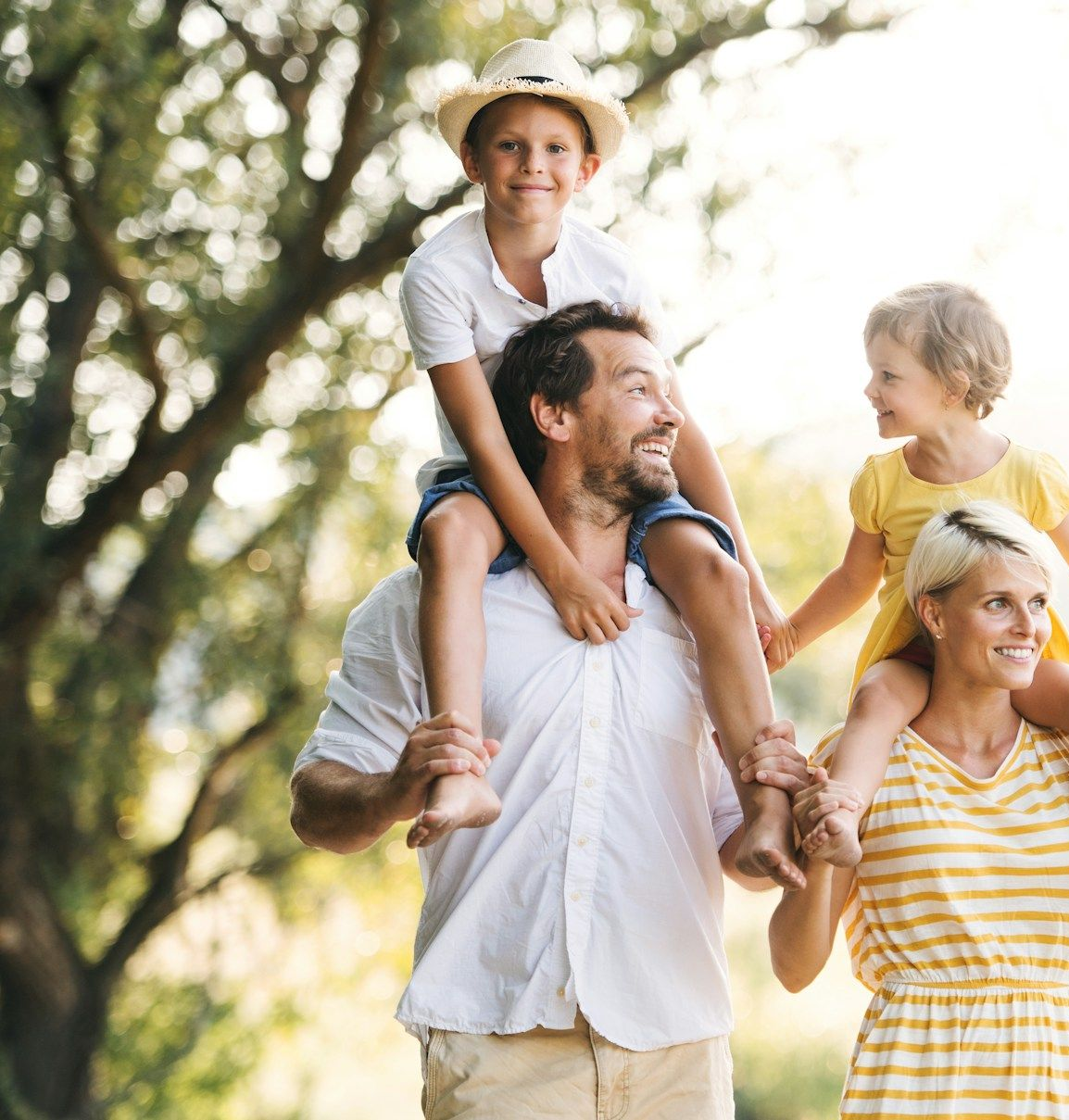 Family outdoors with two children riding on adults’ shoulders in a sunny park