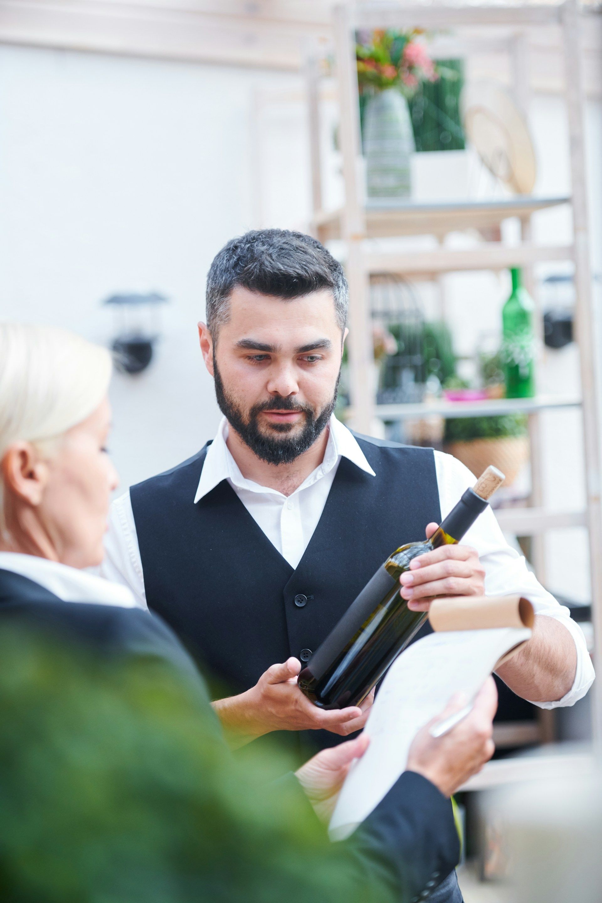 Man in black vest presenting a wine bottle to a customer in a bright shop