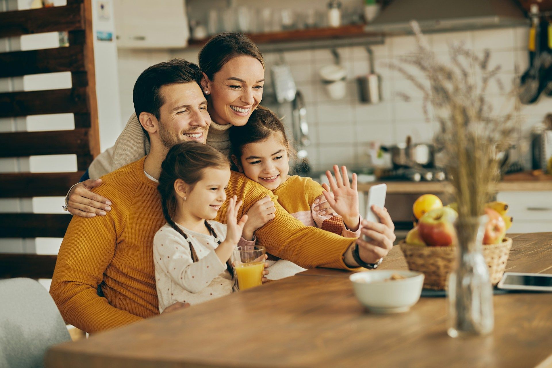 Family of four smiling at a kitchen table while taking a selfie in a cozy home kitchen.