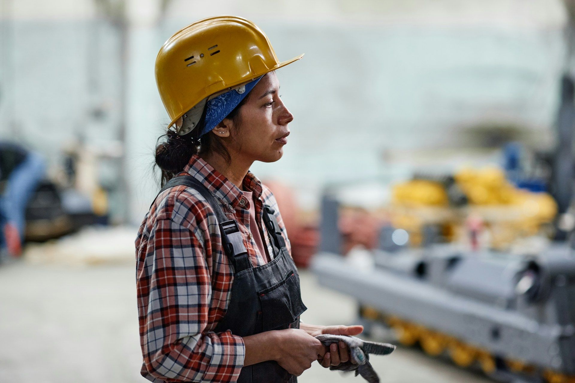 Worker in yellow hard hat and plaid shirt holding a tool in an industrial workshop