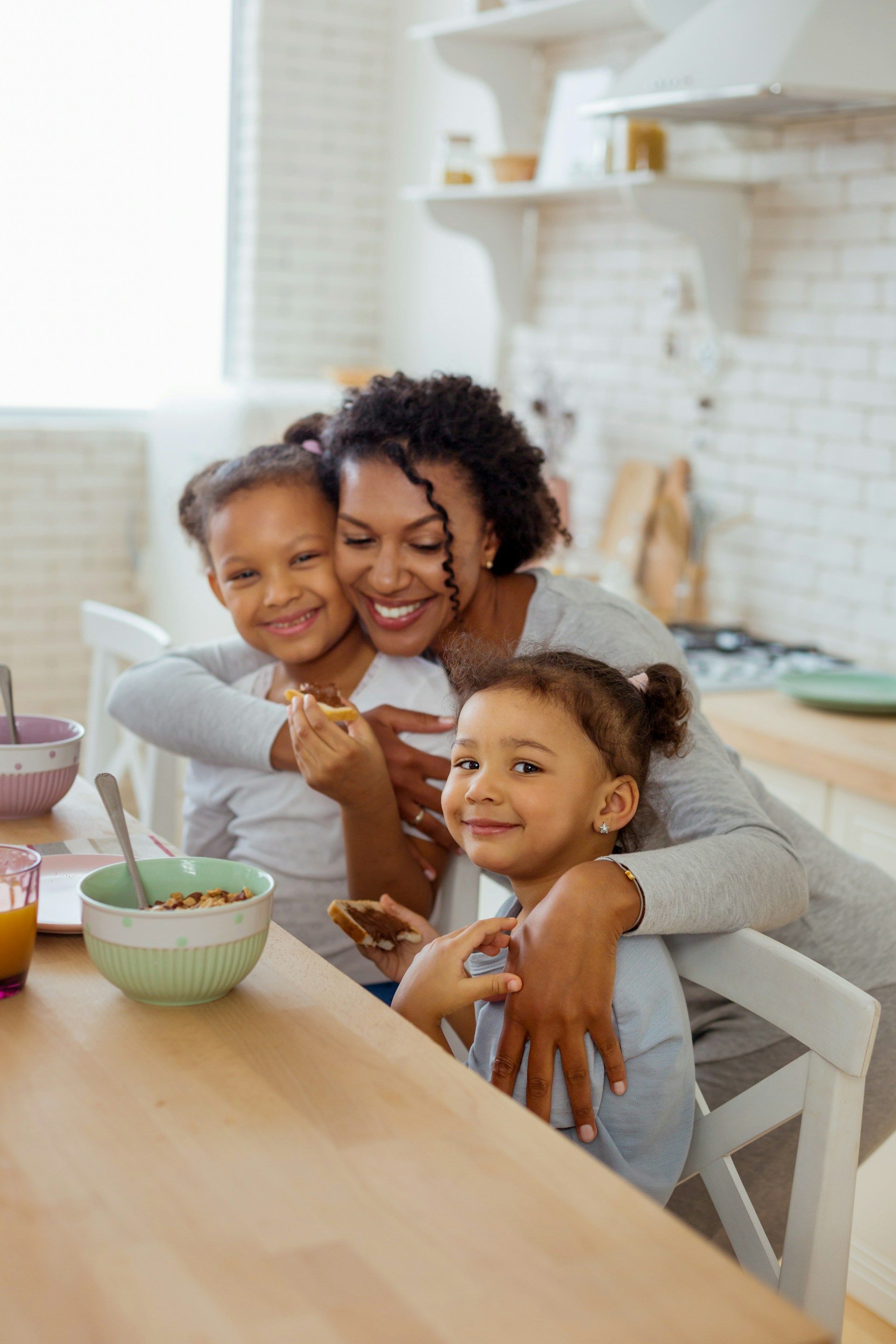 Person smiling with two children at a kitchen table, sharing a snack in a bright home kitchen