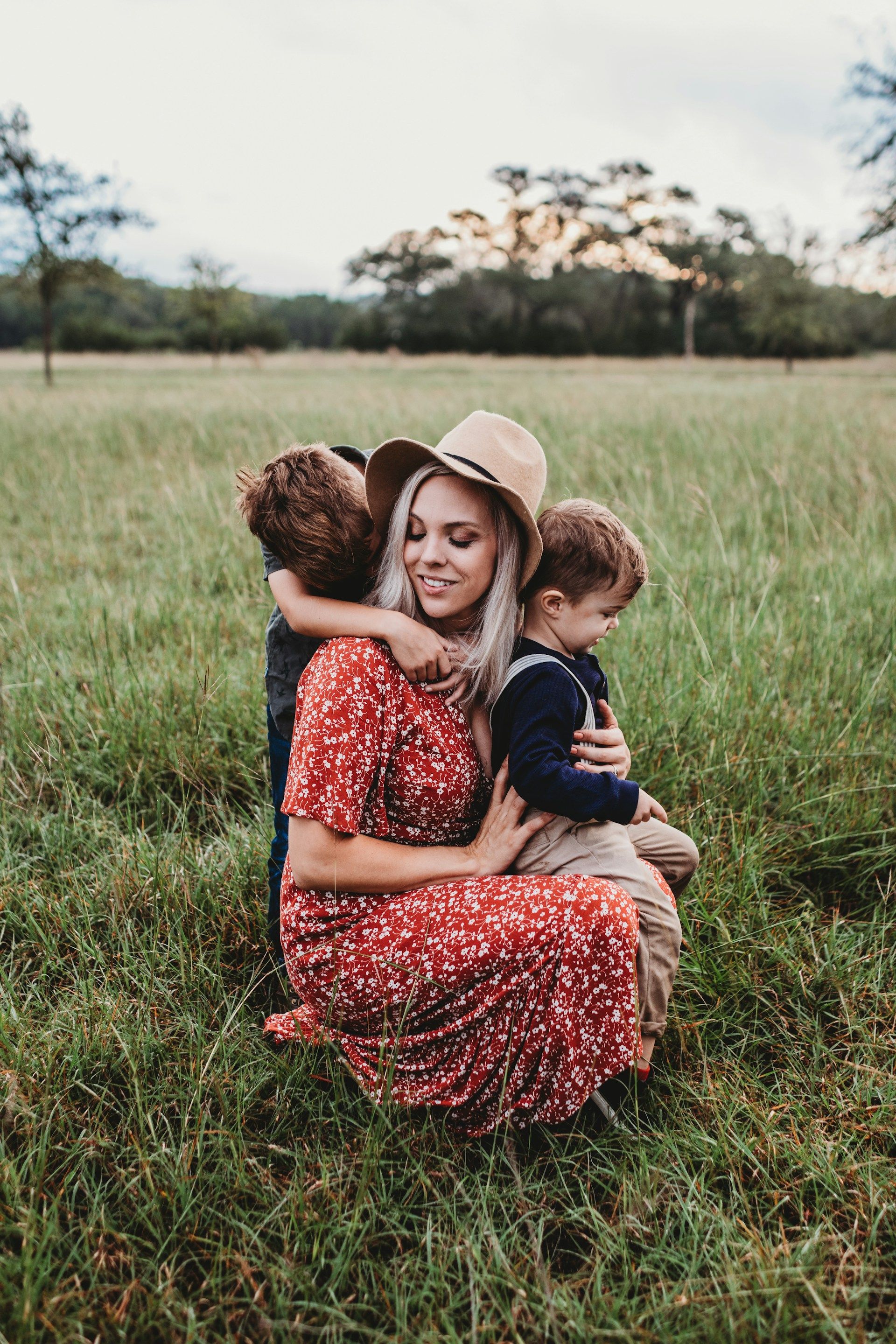 Woman in a red dress and hat hugging two children in a grassy field