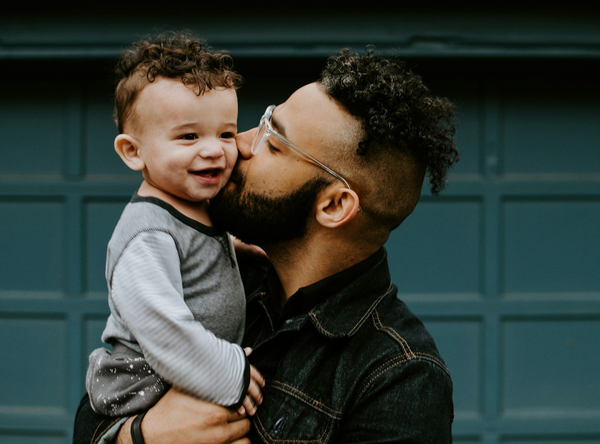 Man kissing a smiling child on the cheek against a dark green garage door backdrop