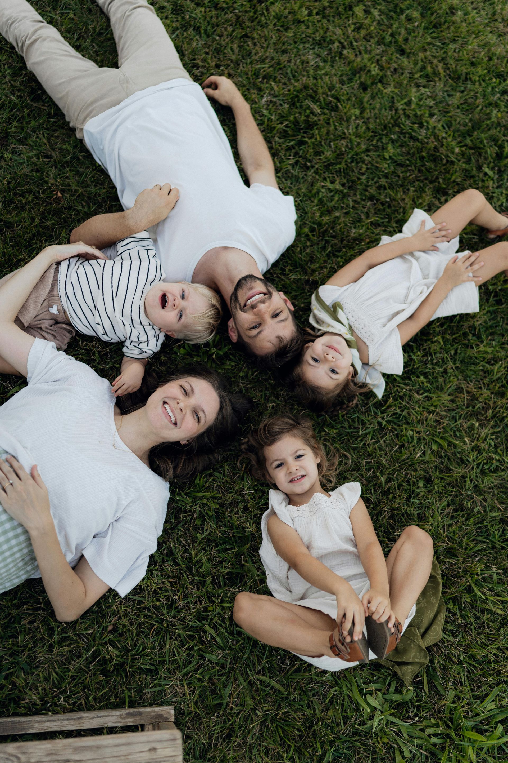 Family lying on grass, smiling up at the camera, with two children and two adults in white clothing.