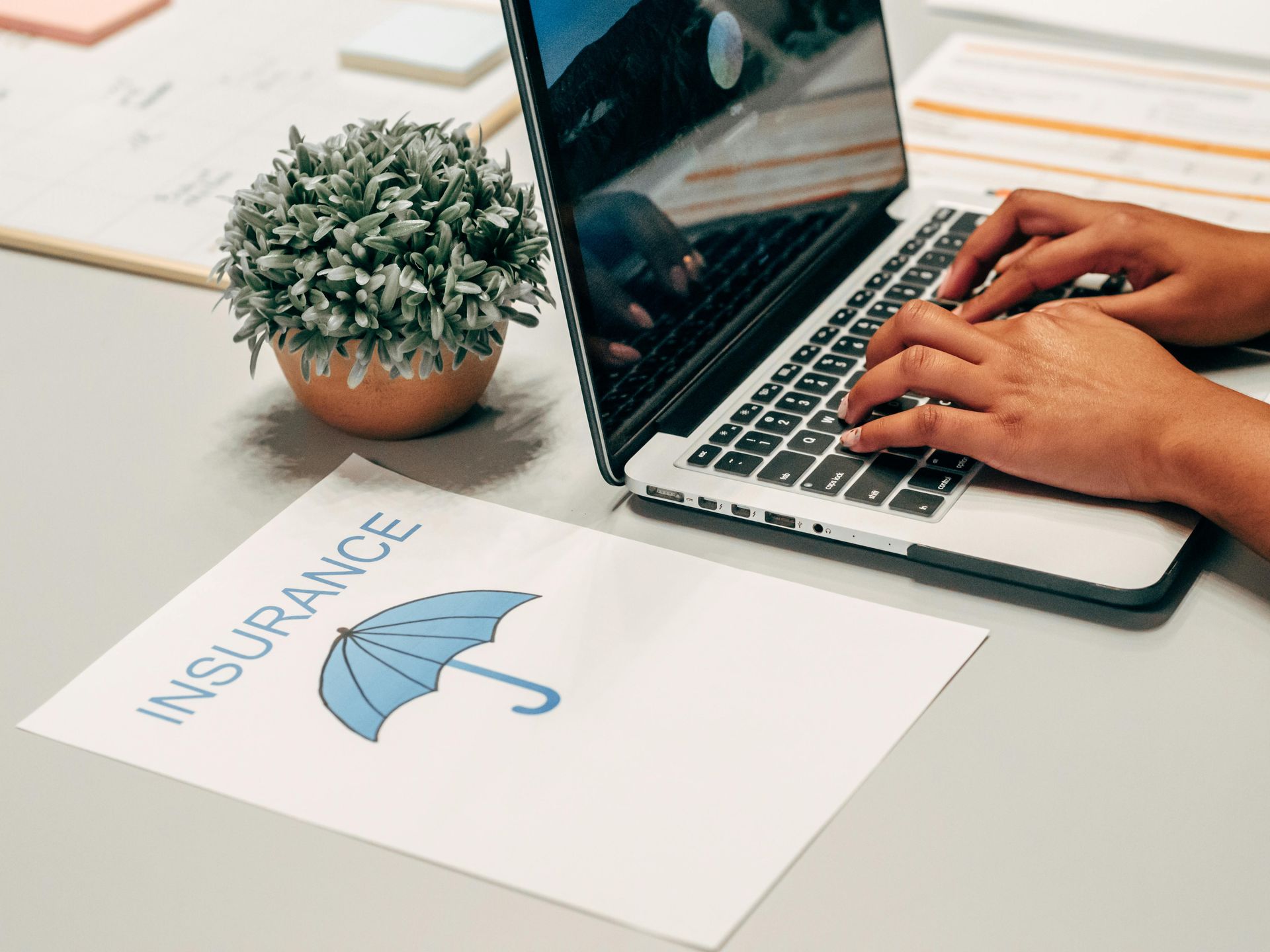 Hands typing on laptop beside insurance document with umbrella icon and potted plant on desk