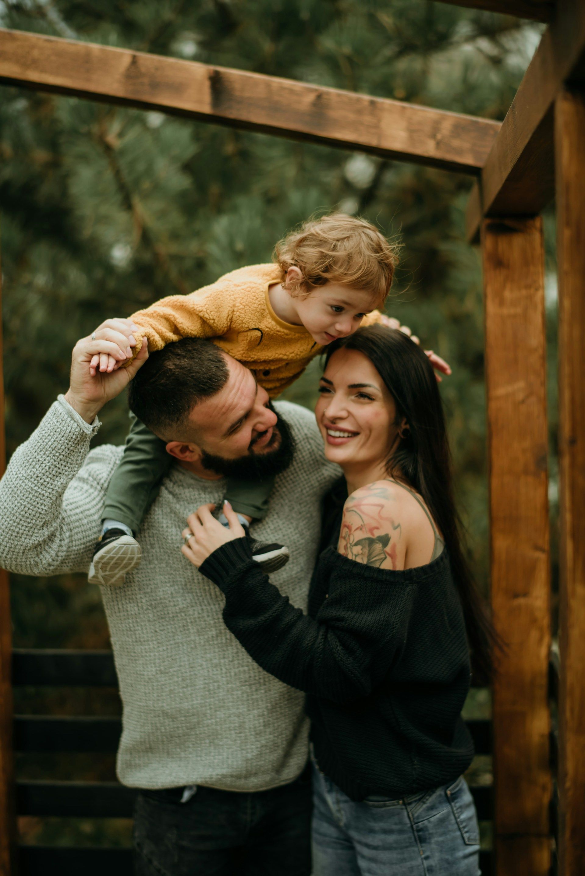Couple and child posing under a wooden arch outdoors, smiling in a warm, wooded setting