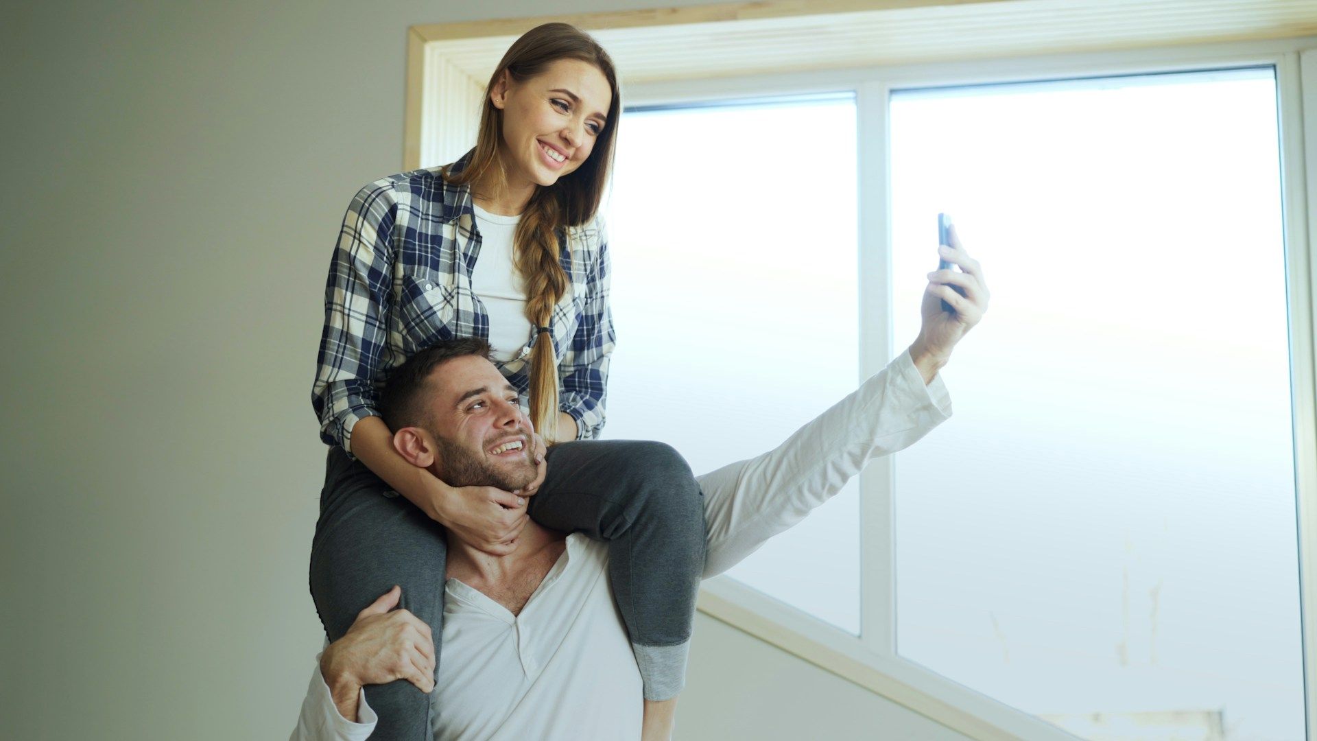 Couple smiling indoors, man taking a selfie with woman on his shoulders near a bright window