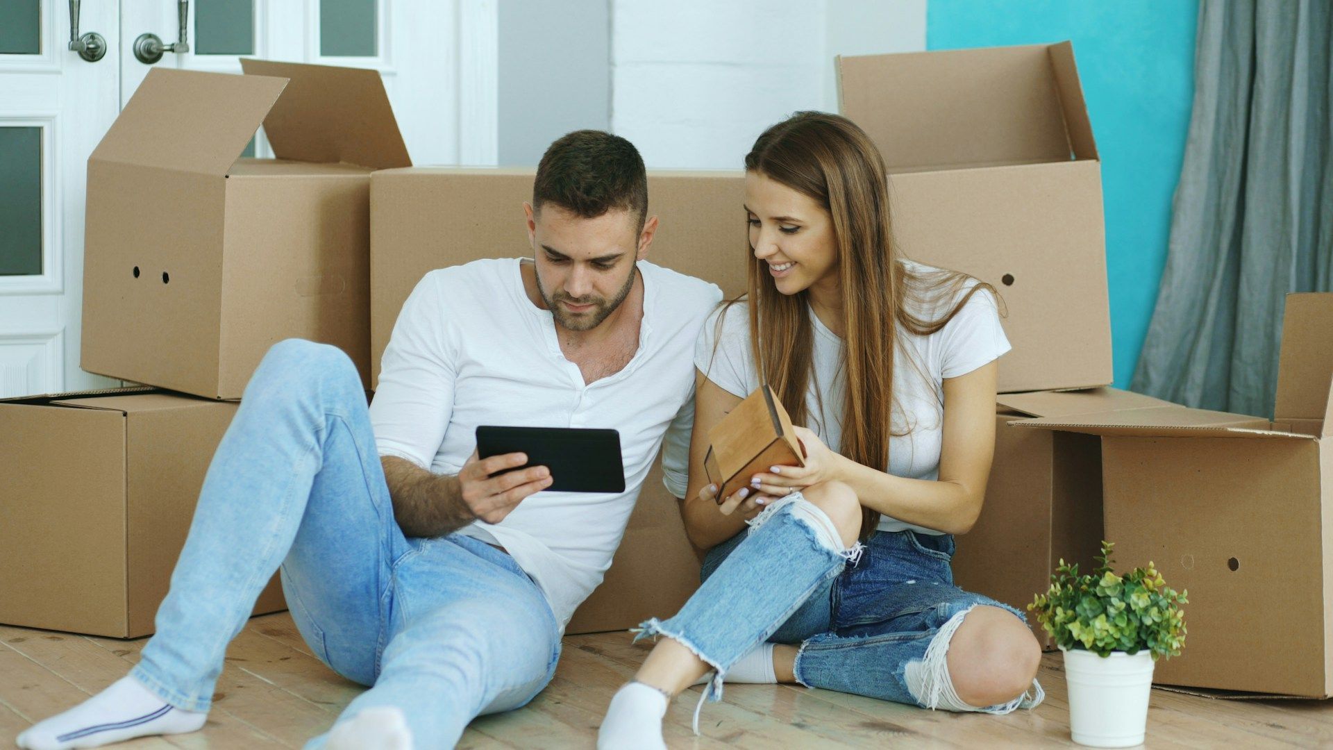 Couple sitting on the floor among moving boxes, looking at a phone in a bright living room.