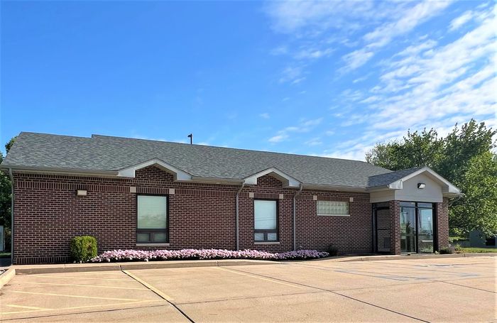Single-story commercial brick building with a gray shingled roof, glass entrance, and a paved parking lot under blue sky.