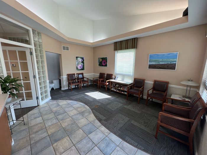 A waiting room featuring rows of brown wooden chairs, tile and carpet flooring, light walls, and decorative wall art.