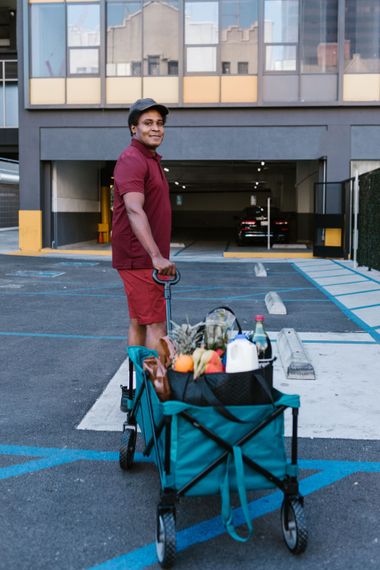 Person standing by a teal cart loaded with supplies in a parking lot outside a building.