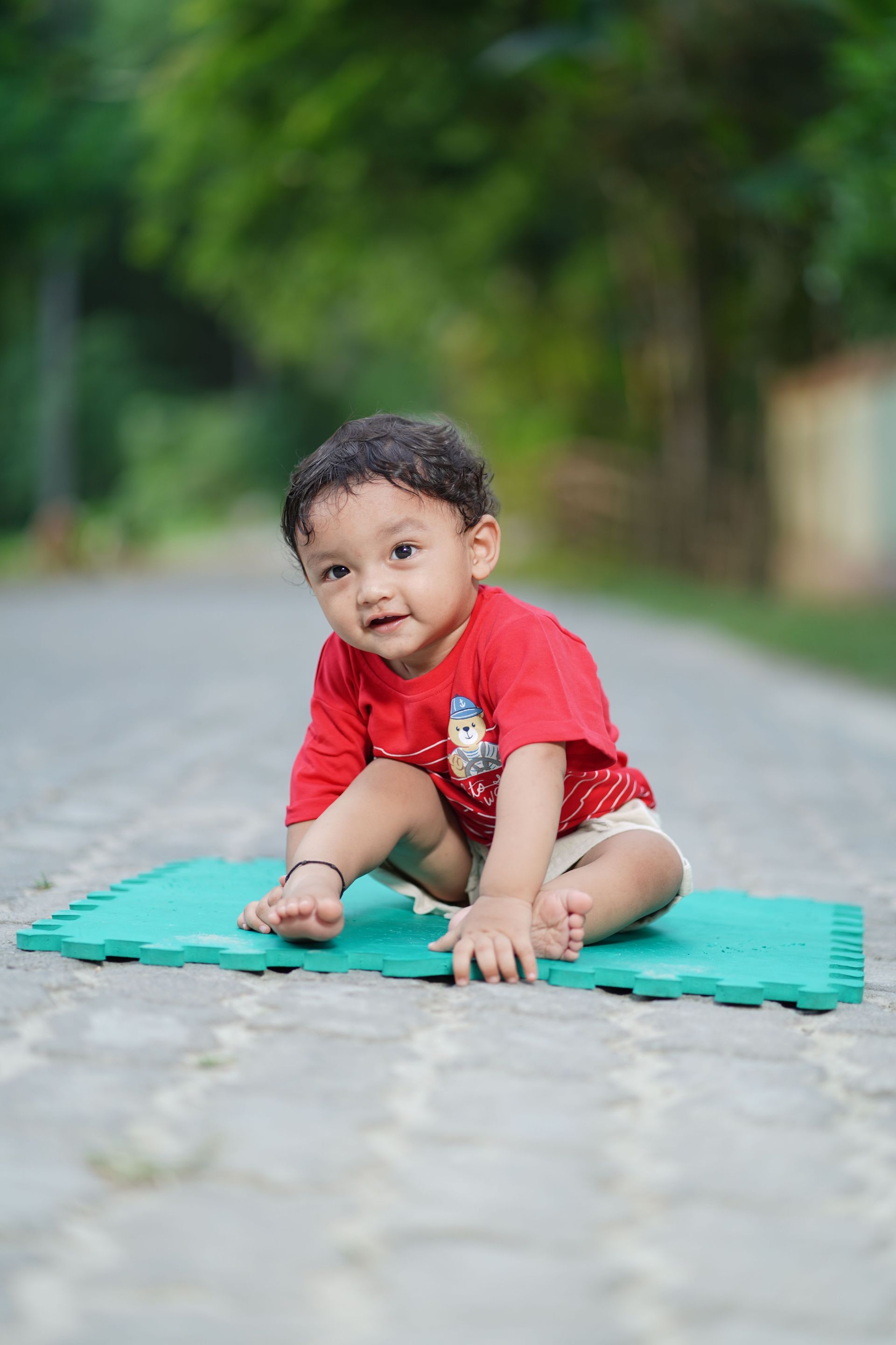 Baby sitting on a green mat on a paved path, wearing a red shirt, outdoors with blurred greenery in the background