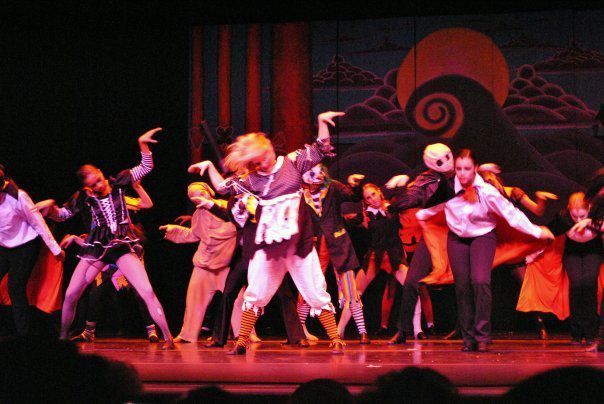 Dancers on a stage perform; a person in a costume center stage is highlighted against a painted backdrop.