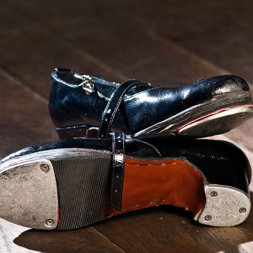 Black tap shoes with silver taps, on a wooden floor. One shoe is stacked on top of the other.