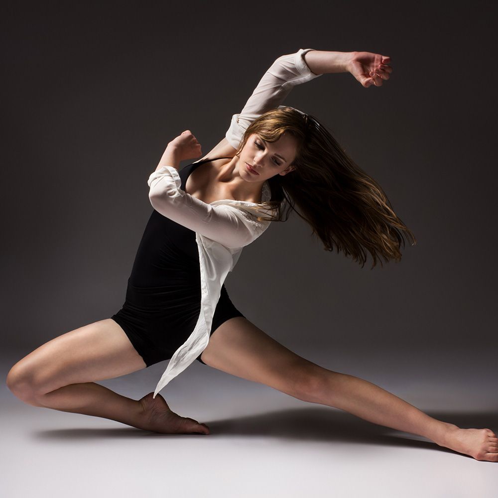 Dancer in a black bodysuit and white shirt, performing a graceful pose with outstretched arms and bent leg.